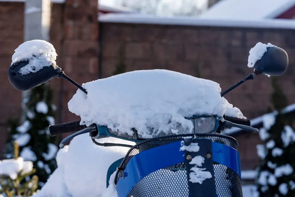 A snow-covered motorcycle sits parked outside with a brick wall and snow-covered shrubs behind it.